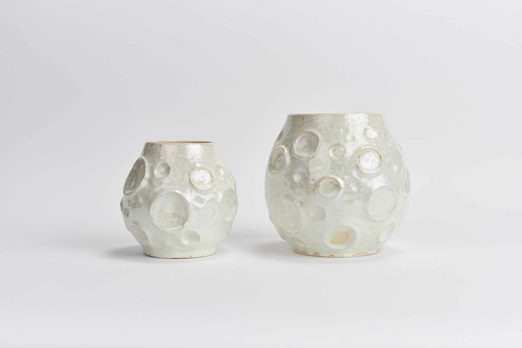 Two porcelain wheel-thrown vases with a textured, pitted surface and raised circular details, resembling moons, shown in a light off-white iridescent glaze on a plain white background.