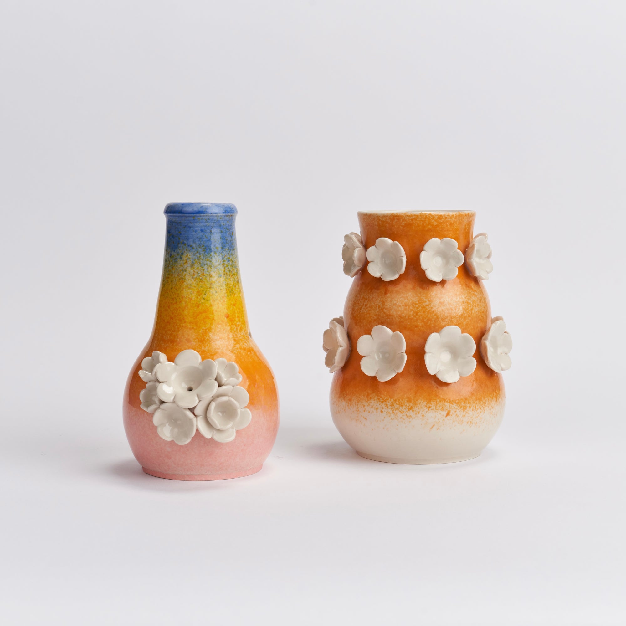 Two wheel-thrown ceramic pieces displayed on a clean white studio background. The left piece is a bubbler (smokeware) and has a narrow neck with a blue-to-yellow-to-orange-to-pink gradient ad a cluster of white flowers. The middle flower is the largest and disguises the bowl. The other piece is a vase with an orange-to-cream gradient and two rows of white flowers sculpted on top.