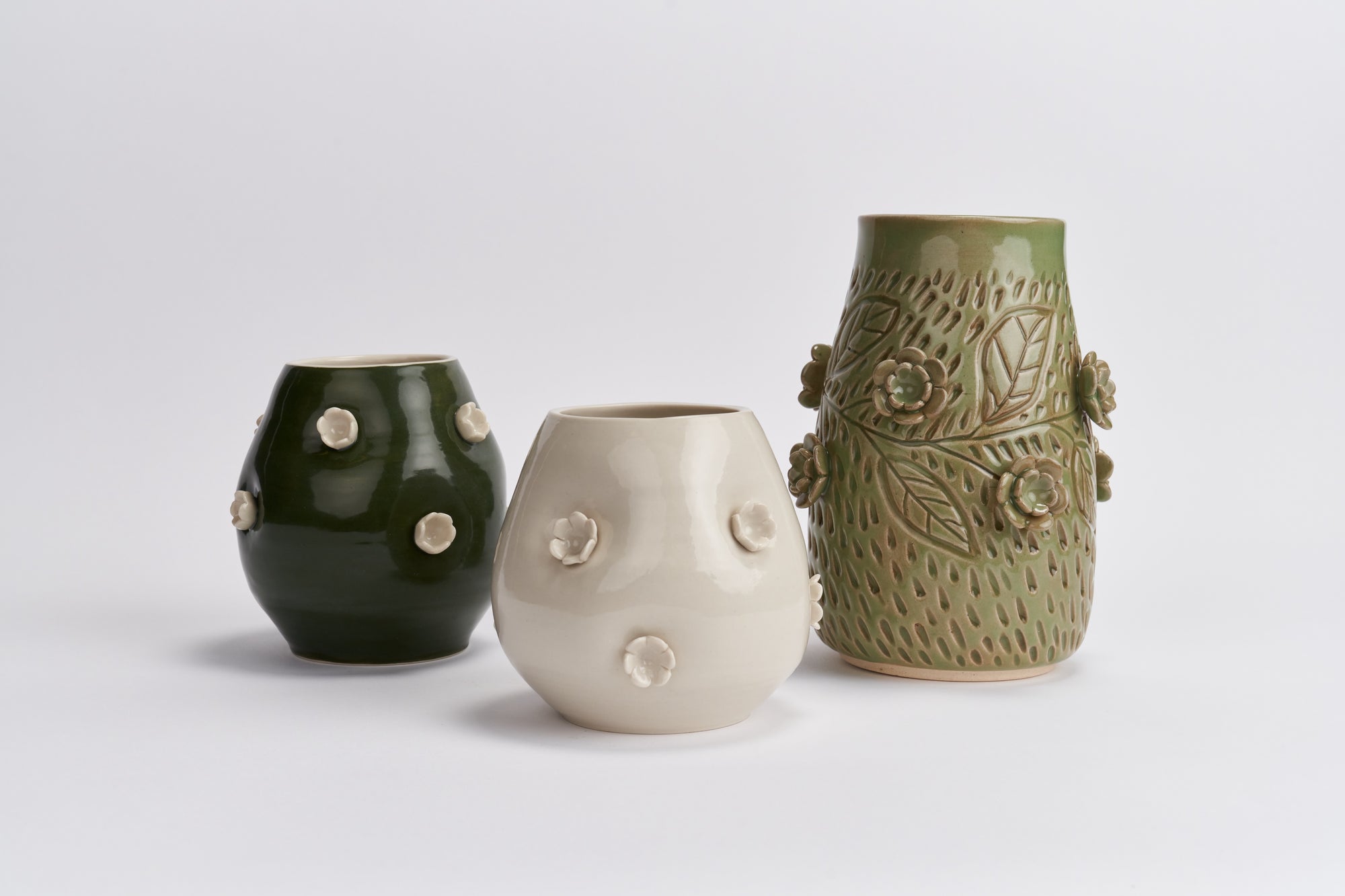Three wheel-thrown ceramic vases with organic shapes and textured glazes, displayed against a neutral white background. The left vase is porcelain, and painted dark green with raised white flowers, the center vase is also porcelain with flowers, covered in a clear glaze, and the right vase is a light stoneware jade green with a leafy embossed pattern and raised floral details.